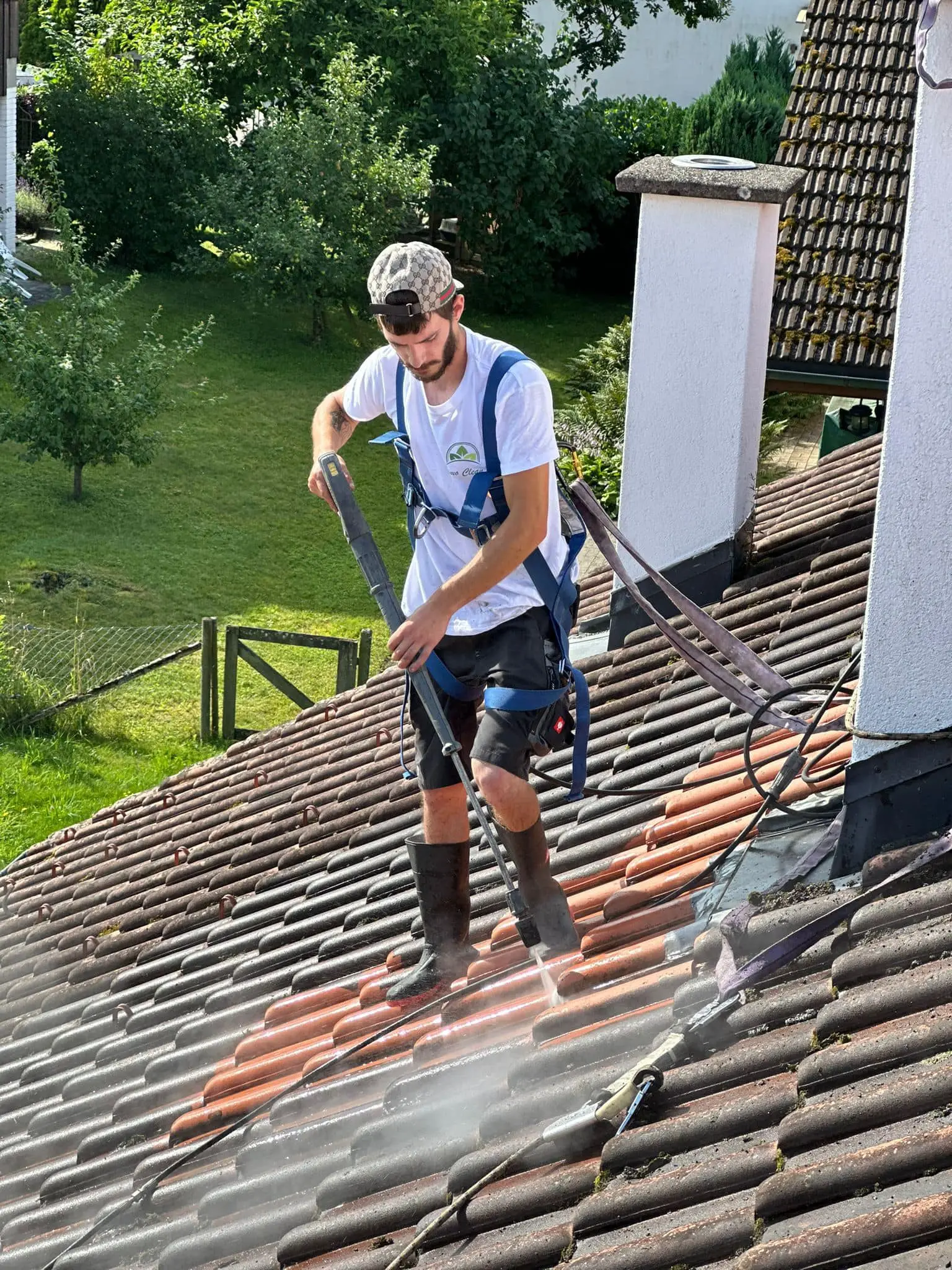 Mitarbeiter von Osmo Clean reinigt gesichert am Gurt ein Ziegeldach, der gereinigte Streifen hebt sich deutlich vom bemoosten Rest ab